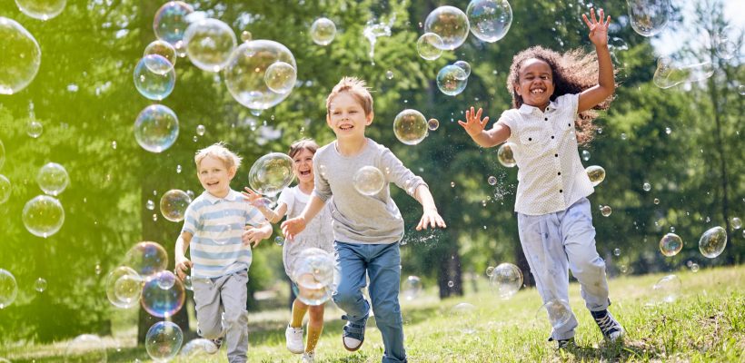 Multi-ethnic group of little friends with toothy smiles on their faces enjoying warm sunny day while participating in soap bubbles show