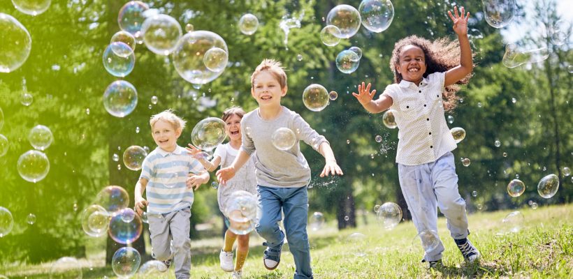 Multi-ethnic group of little friends with toothy smiles on their faces enjoying warm sunny day while participating in soap bubbles show