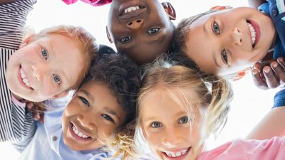 r and smiling at camera. Team of smiling kids embracing together in a circle. Portrait of young boy and pretty girls looking at camera.
